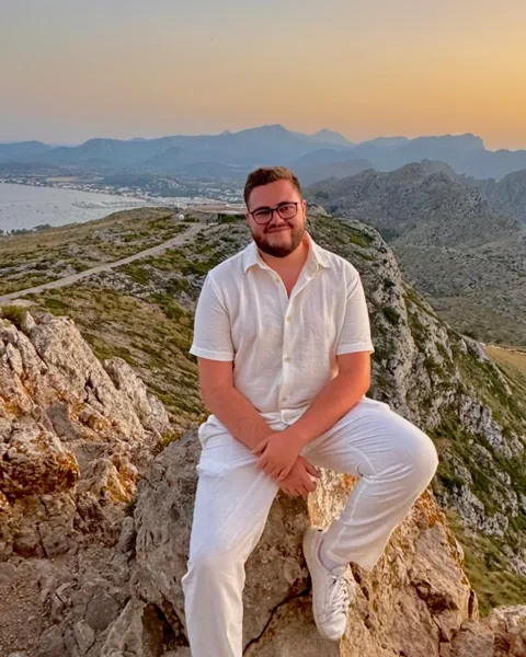Aymeric Mas, souriant, assis sur un rocher face à la mer et aux montagnes au coucher du soleil
